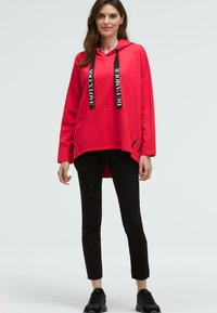 Roter Kapuzenpullover mit schwarzen Kordeln, bedruckt mit Text, lockerer Schnitt und seitlichem Schlitz. Kombiniert mit schwarzen engen Hosen und schwarzen Turnschuhen.