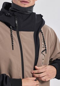 Tarn- und schwarze Jacke mit hohem Kragen, Reißverschluss, verstellbarer Kapuze und Logodetail. Verfügt über strukturierte Stoffe und elastische Bündchen.