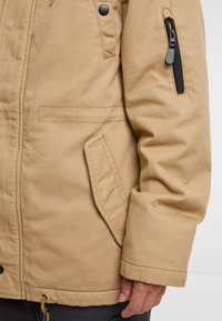 Tan parka jakke med en teksturert finish, som har en glidelåsarm lomme, knappestengte front og sidelommer for funksjonalitet.