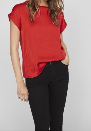 Femme portant un haut rouge à manches courtes amples, légèrement rentrée dans un pantalon noir taille haute, une main dans la poche.