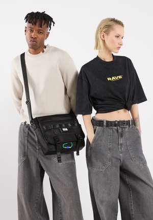 Sac noir à message avec bandoulière réglable, plusieurs compartiments et une accentuation verte. Les mannequins portent des tops oversize et des jeans gris à jambes larges.