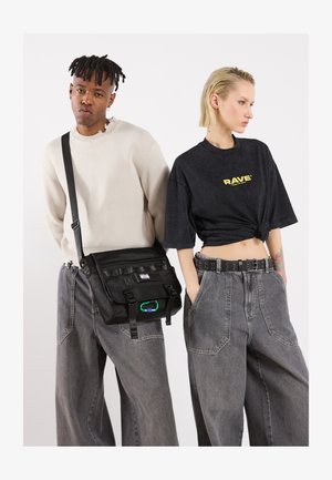 Czarna torba na ramię z regulowanym paskiem, wieloma przegródkami i zielonym akcentem. Manekiny noszą luźne bluzki i szerokie szare dżinsy.