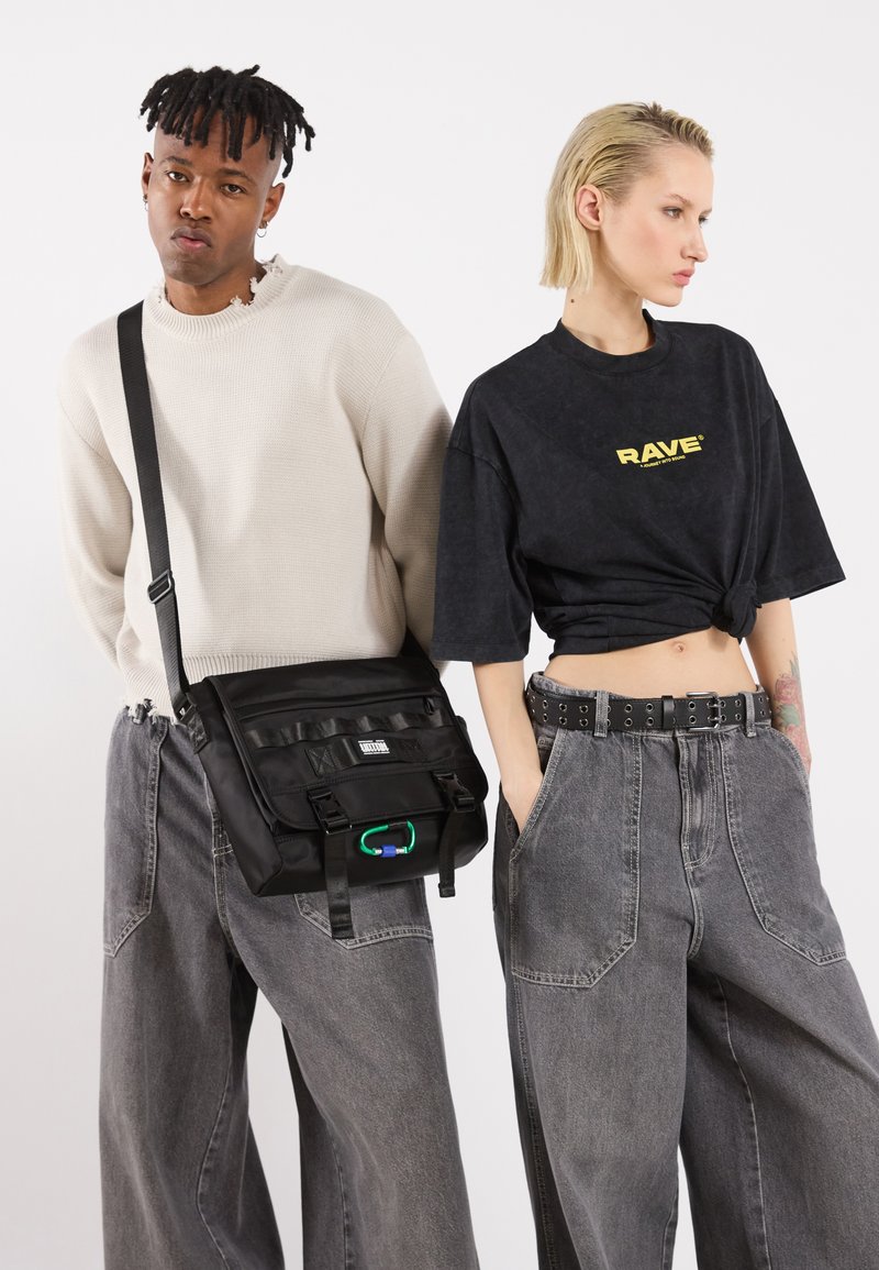 Bolsa de mensajero negra con correa ajustable, múltiples compartimentos y un acento verde. Los modelos llevan camisetas grandes y jeans de pierna ancha grises.