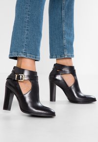 Botines de tacón alto de cuero negro con laterales recortados y hebilla dorada, combinados con jeans azul cortos.