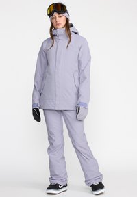 Tenue de ski comprenant une veste imperméable violette clair avec capuche, un pantalon de neige assorti, des baskets noires et des lunettes de ski à verres miroités.