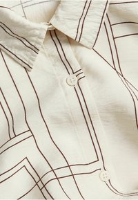 Jasno beżowa koszula z kołnierzykiem, zdobiona brązowymi geometrycznymi liniami oraz z zapięciem na guziki. Wykonana z gładkiego, teksturowanego materiału.