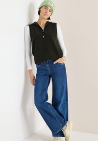 Fleeceweste in Dunkelgrün mit halbem Reißverschluss, kombiniert mit weitem blauen Jeans im Marlene-Stil und beigen Sneakers. Das Modell trägt eine hellgrüne Mütze.