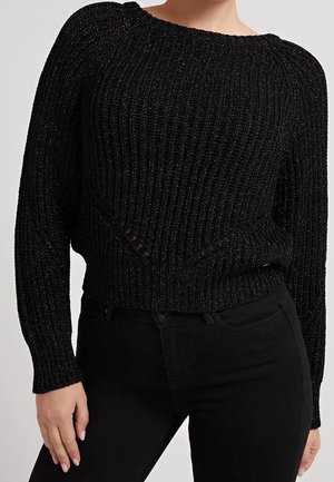 Vrouw draagt een zwart geribd gebreide trui met subtiele glans en zwarte hoge taillebroek, staand met handen ontspannen langs het lichaam.