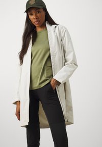 Lätt, lång vit jacka över en grön skjorta med ficka, tillsammans med svarta byxor. En mörkgrön keps fulländar outfiten.
