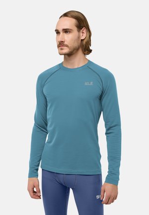 Mann mit schulterlangem Haar, der ein blaues Langarmhemd und dunkelblaue Leggings trägt, steht und schaut zur Seite.