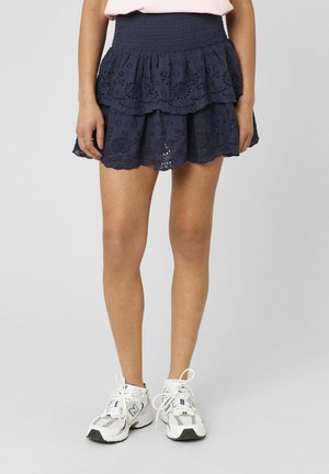 Femme portant une mini-jupe bleu marine à volants en tissu ajouré avec une taille smockée et des baskets blanches New Balance, debout devant un fond uni.