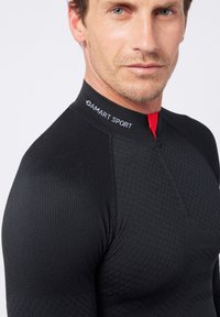 Schwarzes Sportoberteil aus strukturierter Stoff mit hohem Kragen, ausgestattet mit einem Reißverschluss vorne und kontrastierenden roten Details. Logo am Ausschnitt.