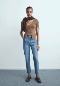 Rjava rebrasta cardigan nad rjavim topom brez rokavov, v kombinaciji s svetlo modrimi ravnimi jeans hlačami in temnimi mokasini. Šal položena okoli vratu.