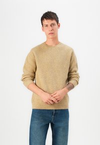 Suéter de malha em castanho claro, com decote redondo, textura canelada e mangas longas, combinado com jeans de denim azuis.