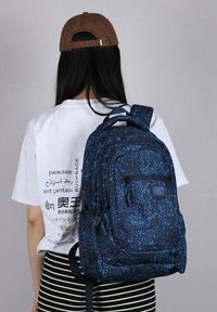 Zaino blu navy con motivo a pixel, dotato di molte cerniere, spallacci imbottiti e una robusta maniglia superiore, indossato da una persona con una camicia bianca e pantaloni a righe.