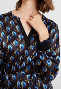 Robe noire à motifs avec des designs circulaires bleus, rouges et dorés. Décolleté en V avec un bord noir et manches bouffantes avec des poignets froncés.