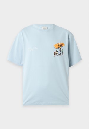 Uomo in piedi che indossa una maglietta azzurro chiaro con grafica di un autobus scolastico giallo e persone, pantaloni neri larghi e scarpe beige su sfondo semplice.