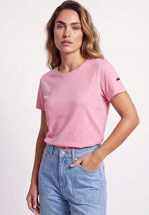 Femme portant un t-shirt rose à manches courtes et un jean taille haute bleu clair, debout avec une main dans la poche devant un fond uni.