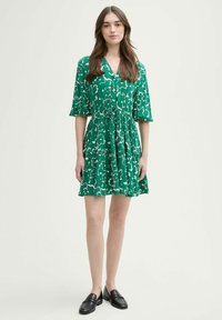 Vestido verde con un patrón abstracto blanco, mangas cortas abullonadas y cintura fruncida. Combinado con mocasines negros. Tejido suave, diseño casual.