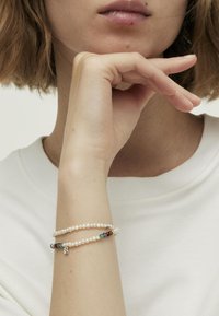 Perłowa bransoletka z kolorowymi koralikami, warstwowy design, z małym charms'em. Noszona na nadgarstku, na tle jasnej koszuli.