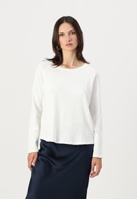 Woman with straight dark hair wears white long-sleeve top and navy blue satin skirt, standing against a light background.