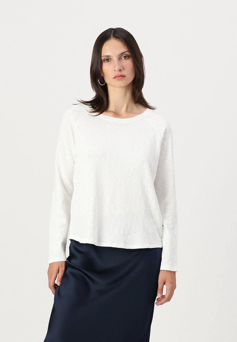 Woman with straight dark hair wears white long-sleeve top and navy blue satin skirt, standing against a light background.
