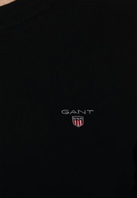 Schwarzer Strickpullover mit Rundhalsausschnitt, geripptem Muster und einem kleinen bestickten GANT-Logo mit Schild-Design auf der Brust.