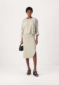 Top beige chiaro con maniche ampia, sovrapposto a una gonna midi aderente. Sandali neri con tacco a blocco e una piccola borsa nera completano il look.
