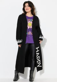 Long cardigan noir avec des flammes blanches sur les manches et le texte "HAPPY". Porté sur un t-shirt graphique violet et des leggings noirs avec des bottes cloutées.
