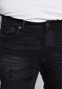 Los jeans de mezclilla negra presentan un corte ajustado con una textura suave, un ligero desgaste y detalles intencionadamente rasgados en el muslo.