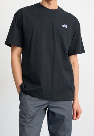 Persona che indossa una t-shirt nera ampia con un piccolo logo di sneaker sul petto e pantaloni grigi, in piedi con una mano in tasca.