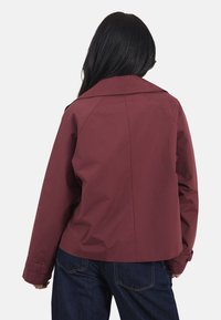 Veste légère bordeaux avec un col en notch, une couture centrale dans le dos et des poignets boutonnés. La texture visible est lisse et mate.
