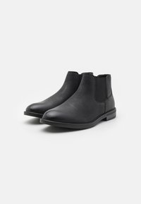 Las botas de tobillo de cuero negro cuentan con una punta redonda, paneles laterales elásticos y un tacón bajo apilado. La textura es suave con un grano sutil.