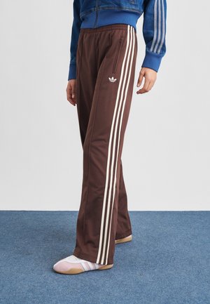 Personne portant un pantalon de survêtement Adidas marron à bandes blanches, une veste bleue avec des bandes assorties, et des chaussettes blanches et roses sur une moquette bleue.