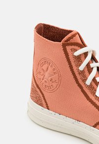 Sapatilha de cano alto em malha coral com atacadores brancos. Apresenta detalhes texturizados nas laterais e um logótipo circular. Sola de borracha com design salpicado.