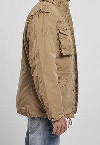 Mężczyzna w beżowej kurtce roboczej z wieloma kieszeniami i dżinsach, pokazany z boku na jednolitym tle.