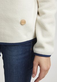 Fleecasti pulover u krem boji s tamnoplavim obrubom i drvenim logotipom. Ima meku, teksturiranu površinu i opušteni kroj.