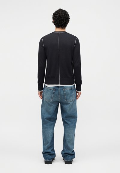 Homme aux cheveux bouclés portant un haut noir à manches longues avec des coutures blanches et un jean bleu large, debout, de dos sur un fond uni.