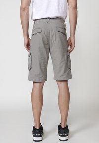 Shorts cargo gris avec poches latérales, passants de ceinture et coupe décontractée. Portés avec des baskets noires présentant des accents blancs. Texture minimale.