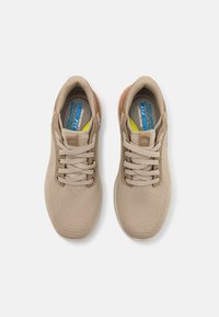 Zapatillas deportivas beige con parte superior de malla, cordones planos y suela texturizada. El interior presenta una etiqueta de plantilla en azul y amarillo.
