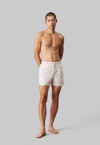 Shorts de bain blancs avec une taille élastique et un cordon de serrage, fabriqués à partir d'un matériau léger, stylés avec une coupe décontractée, arrivant au genou.