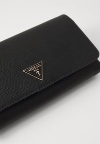 Portefeuille texturé noir avec un matériel doré en forme de triangle portant le logo "GUESS", fermé par un rabat.