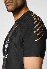 Μαύρο αθλητικό μπλουζάκι με υφή υφάσματος, χρυσές γεωμετρικές λεπτομέρειες στον ώμο και λογότυπο στο μπροστινό μέρος. Κοντινή εικόνα.