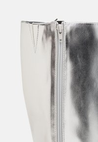 Stříbrná kovová kotníková bota s hladkým koženým povrchem, středovým zipem a strukturovaným tvarem se špičatým horním okrajem.