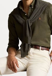 Olijfgroene button-up shirt gecombineerd met een donkergrijze gebreide trui om de schouders geknoopt, beige broeken en een tan leren riem.