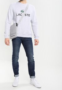 Felpa grigia in cotone con logo "LACOSTE", jeans scuri in denim e sneakers bianche. Borsa a tracolla grigia con il marchio "Calvin Klein".