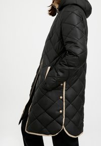 Sort quiltet dunjakke med hætte, beige kant, sidelommer og trykknapper. Har en knælængde pasform og glat tekstur.