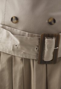 Tela de gabardina beige con dos botones marrones, un cinturón ancho con una hebilla rectangular de carey y pliegues cosidos debajo.