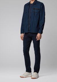Man som bär en mörkblå denimjacka över en marinblå skjorta, mörkblå jeans och vita sneakers med gummisulor, står mot en plain ljus bakgrund.