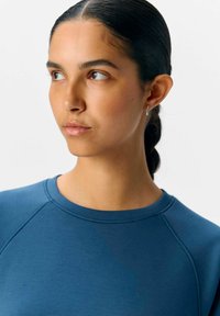 Blaues Sportshirt mit Rundhalsausschnitt und Raglanärmeln, aus glattem Stoff. Das Modell hat glattes Haar und minimale Accessoires.